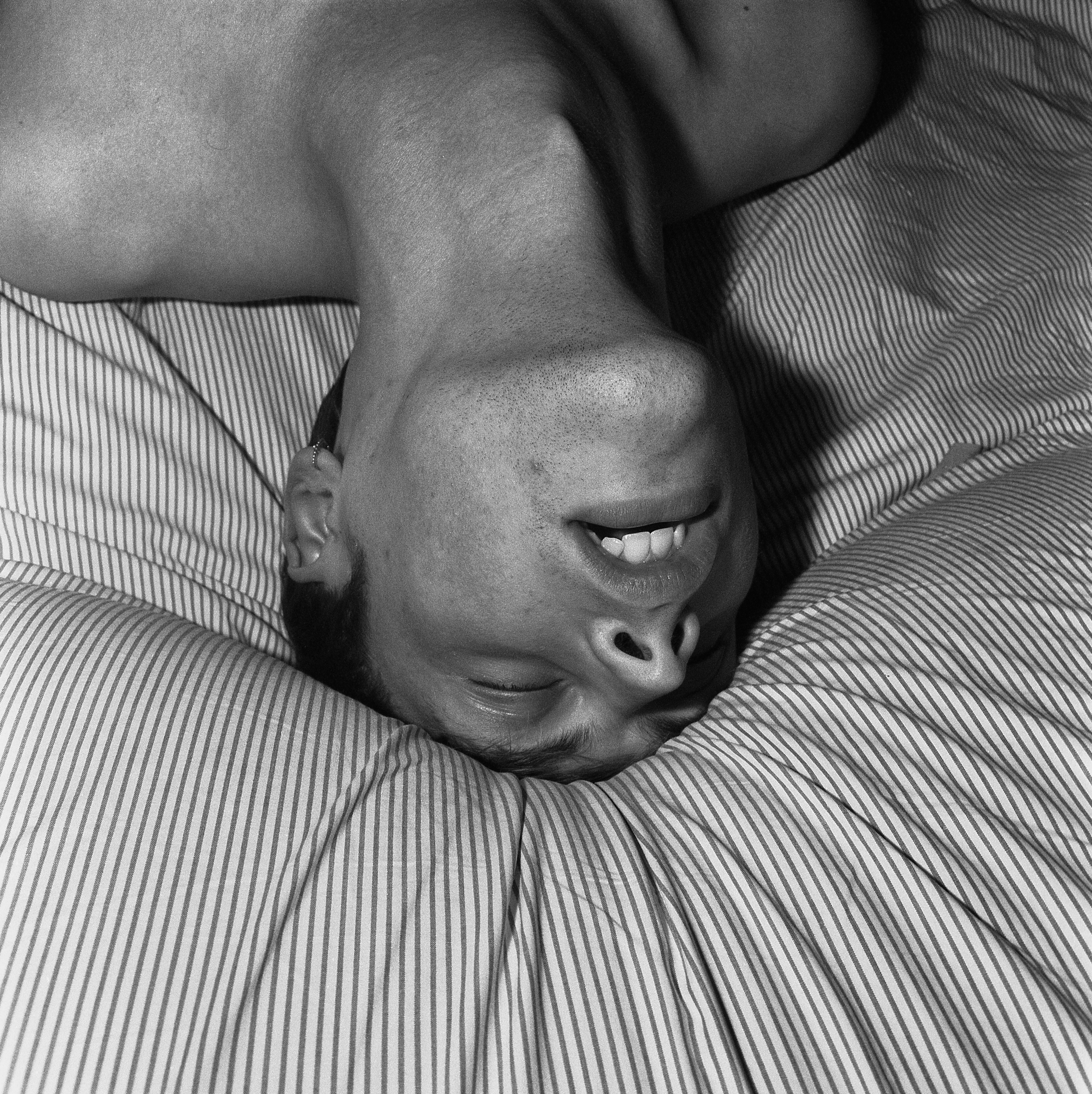 Upside-down photo of a person with short hair arching their back and pushing the top of their head into the bed, their mouth open.