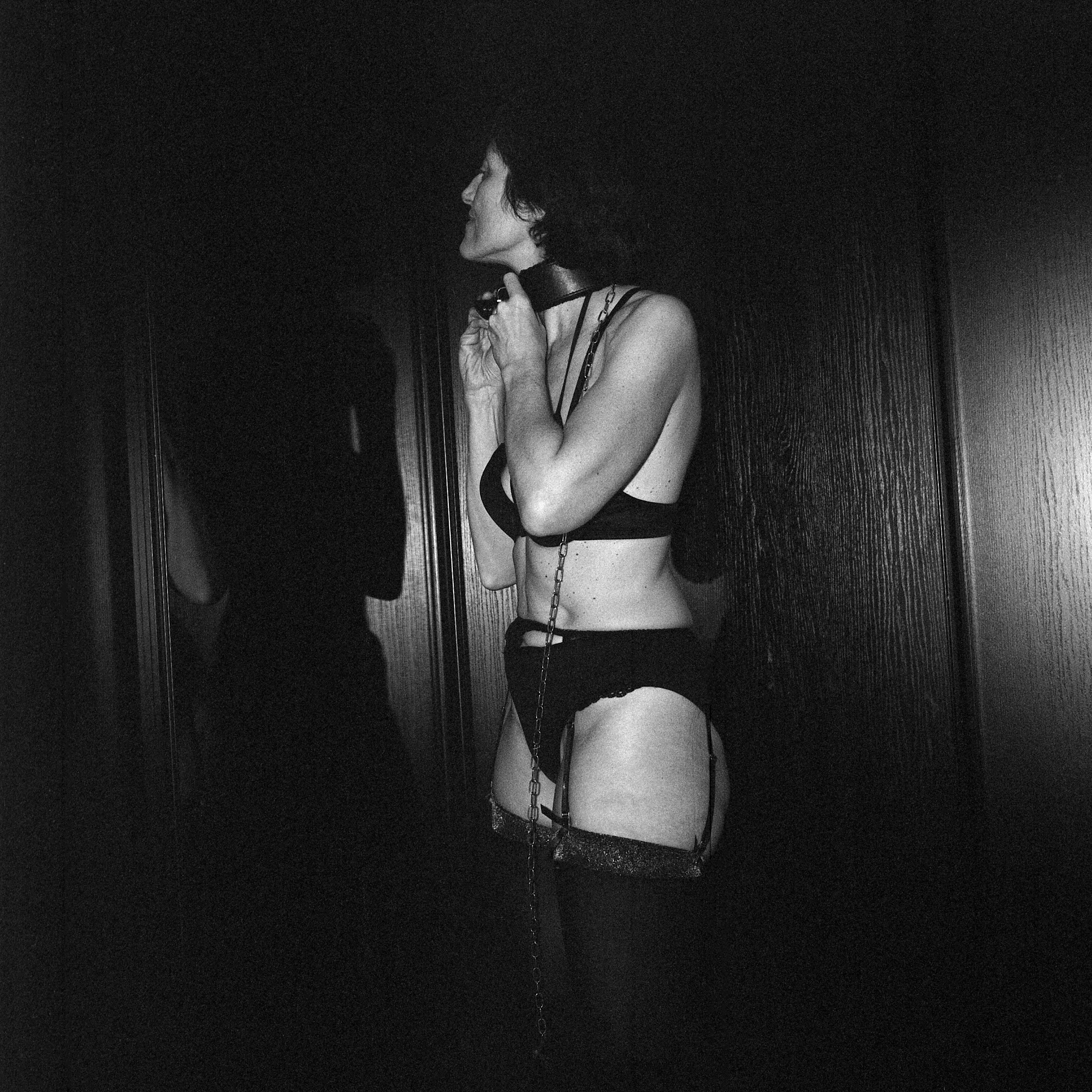 Photo of a middle-aged woman in lingerie and a collar with a chain, standing in a room, her shadow projected onto the wall
