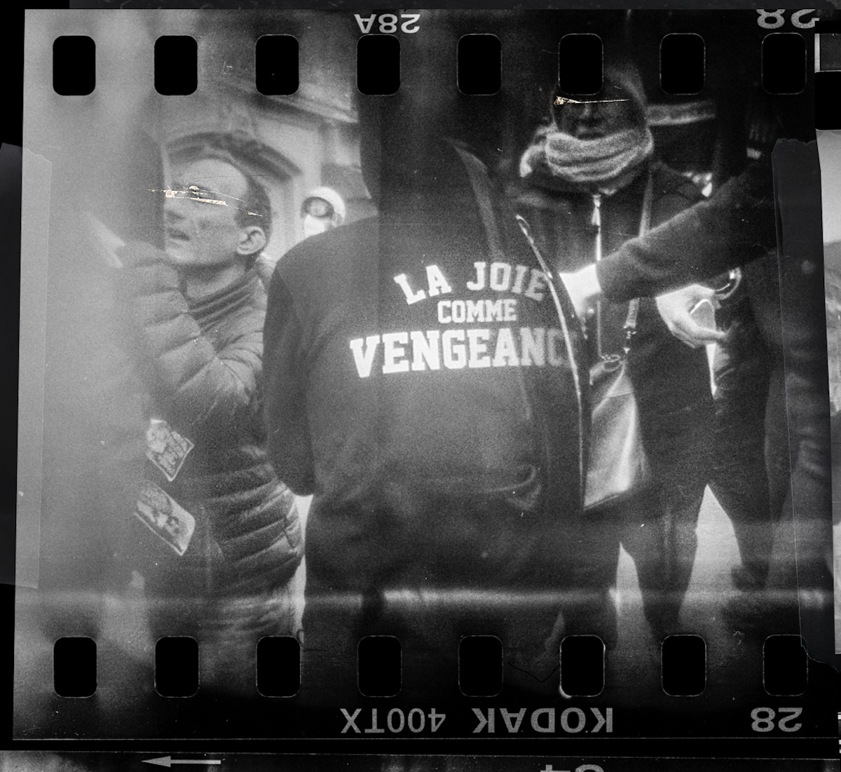 Steven Monteau photos with police weapons: black and white negative of a people at a protest, one is weariig a jumper saying: joy as revenge