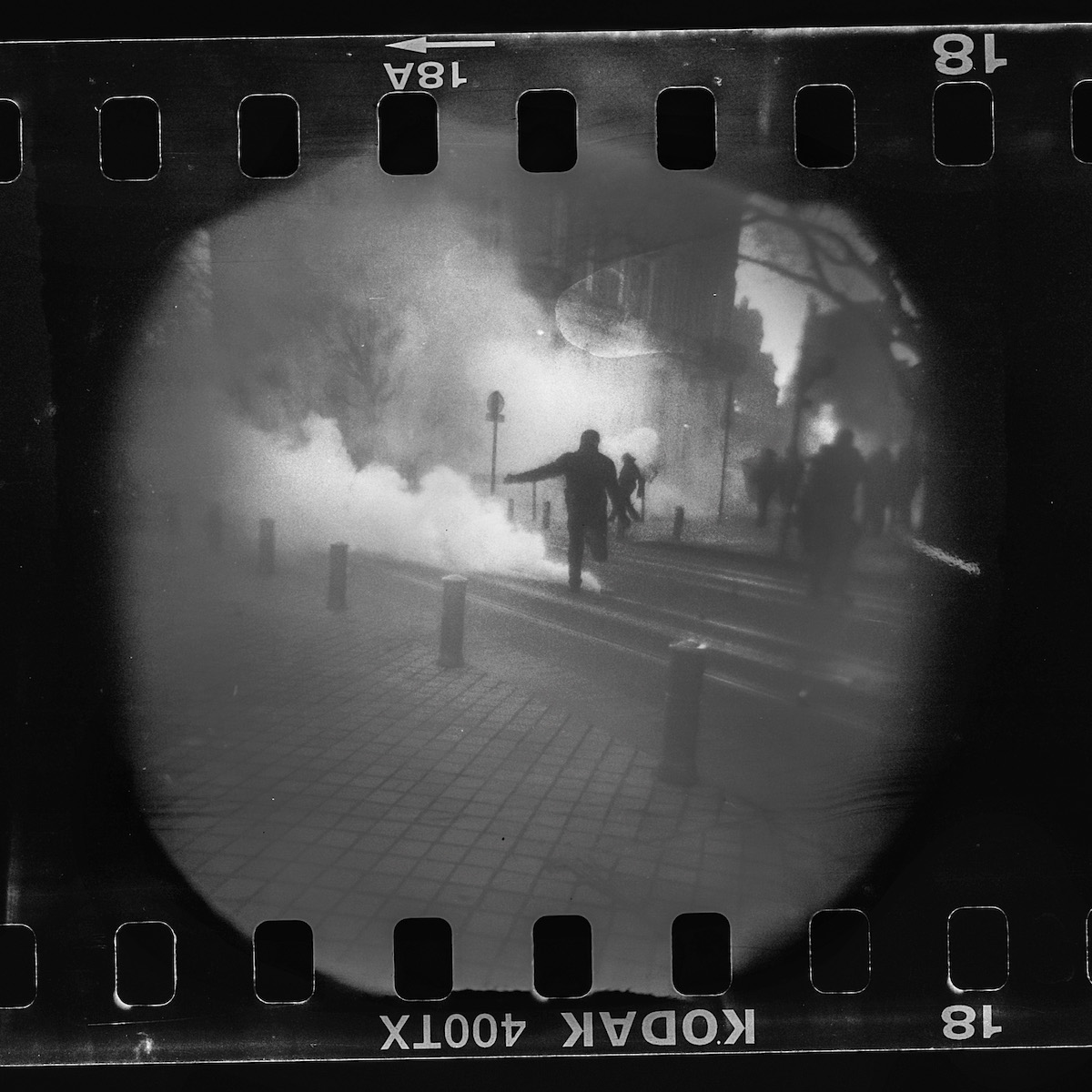 Steven Monteau photos with police weapons: black and white negative of a riot, with smoke and people running on the streets