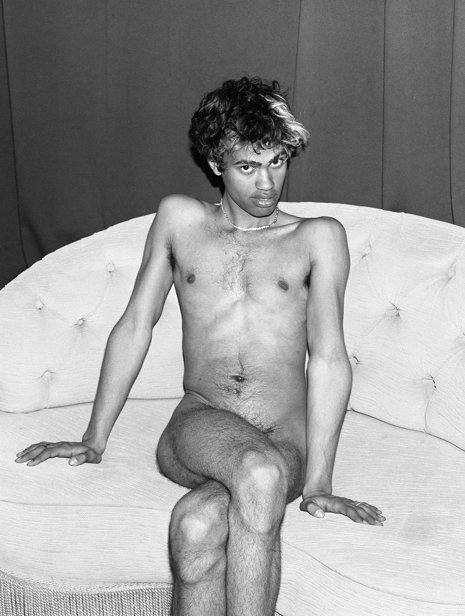A black and white full body photo of a naked person sitting on a vintage couch. They wear a necklace and have a light strand of hair