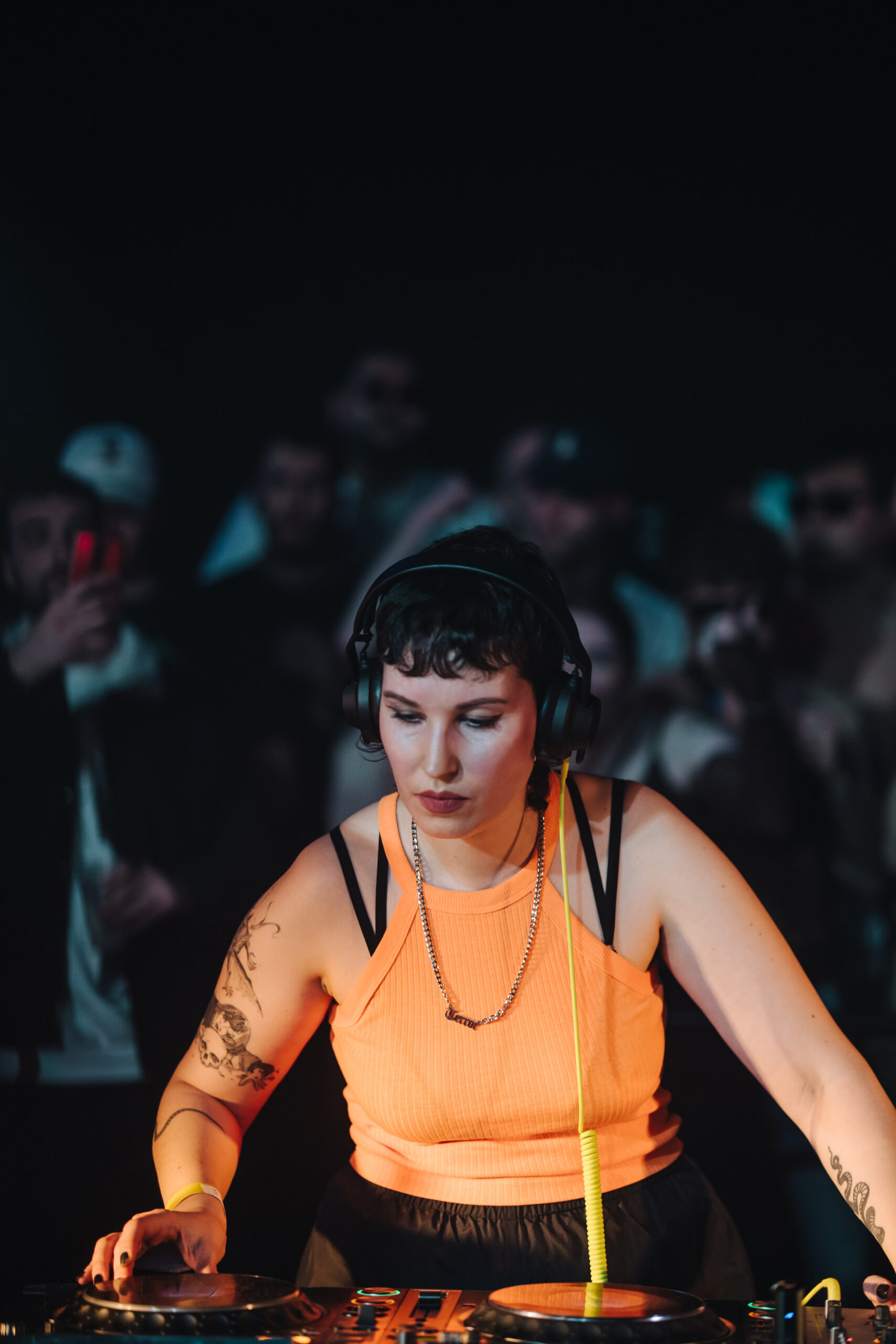 Eine DJ steht in orangenem Top und mit Kopfhöreren vor CDJs