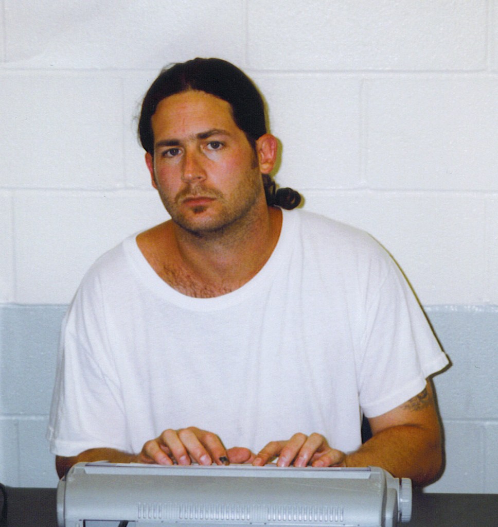 A man with a pony tail and a white T shirt poses in front of a typewriter in prison
