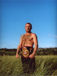 A topless middle aged white man poses with his bare knuckle fighting belt in front of a clear blue sky