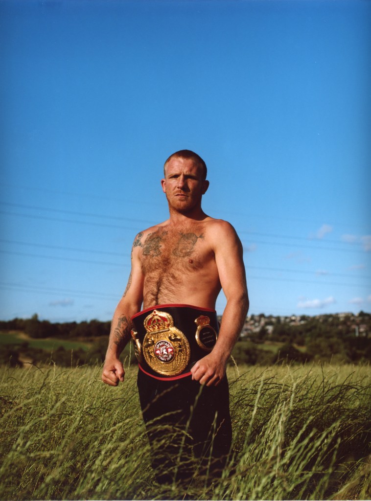 A topless middle aged white man poses with his bare knuckle fighting belt in front of a clear blue sky