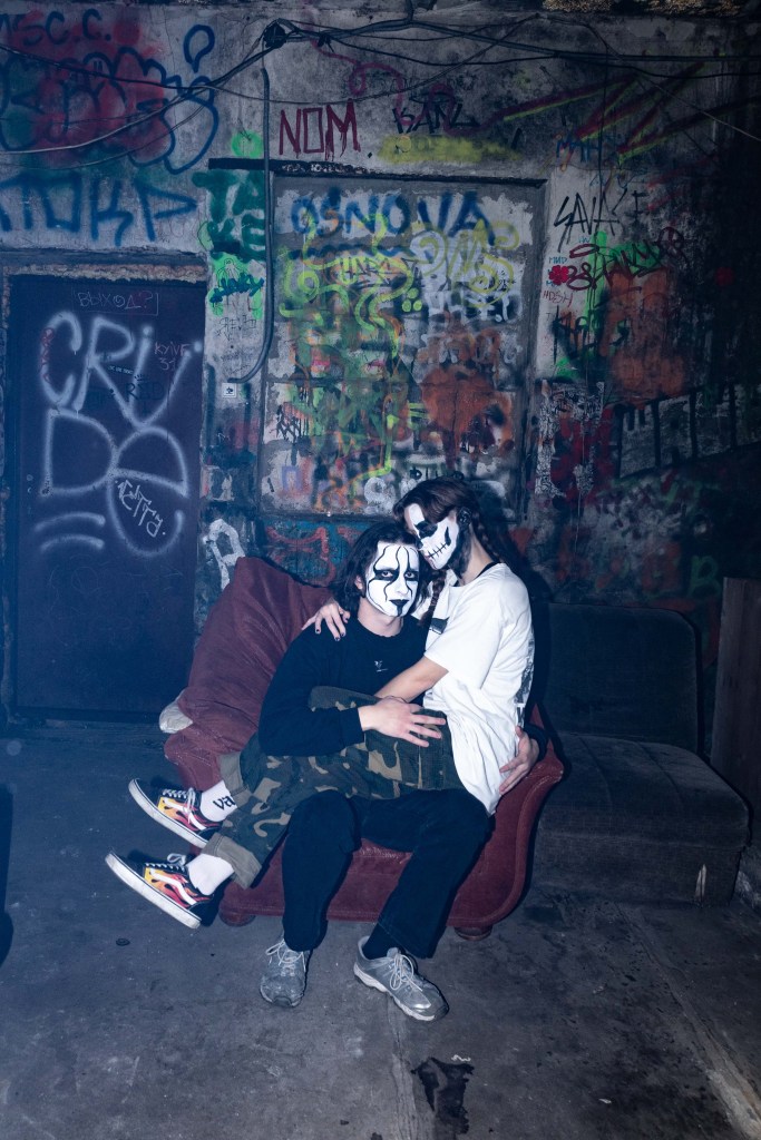 Two goths sit together on a chair in a derelict building